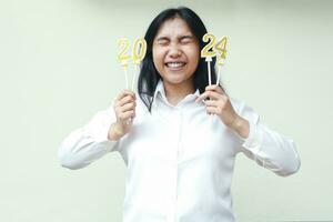 beautiful asian young woman excited for 2024 new years celebration by holding golden numbers candles and close eyes wear white shirt formal isolated photo