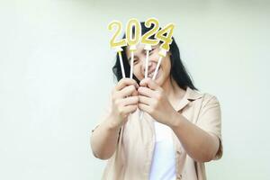 emocionado joven mujer asiático demostración oro velas números 2024 sostener en mano con contento expresión a celebrar nuevo años víspera vistiendo marrón camisa aislado en blanco foto