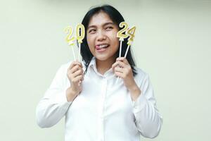 portrait of excited asian young female office worker thinking resolution idea to celebrate 2024 new years eve by holding numeral golden candles on hand wearing white formal suit isolated photo