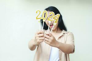 excited young woman asian showing golden candles numbers 2024 on hand to celebrating new years eve wearing casual brown clothes isolated by white background with copy space photo
