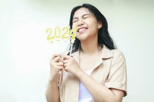 asiático joven mujer emocionado a celebrando nuevo años víspera con participación dorado velas números 2024 y cerrado ojo vistiendo casual marrón camisa aislado por blanco antecedentes foto