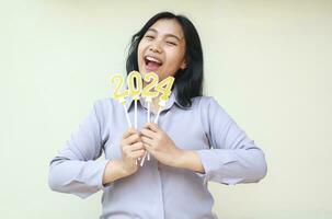 portrait of excited young asian business woman celebrating new years eve with holding candles numbers 2024 and raising her hand wearing grey shirt isolated photo