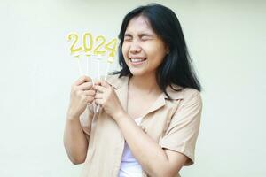 excited asian young woman celebrating new years eve by holding golden candles numbers 2024 with closed eye wearing casual brown shirt isolated on white background photo