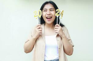 portrait of excited asian young woman scream holding gold candles numbers 2024 to celebrating new years eve wearing casual brown shirt looking away isolated on white background photo