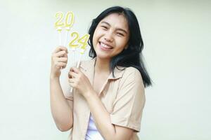 excited asian young woman smiling and holding golden candles numbers 2024 to celebrate new years eve wearing casual brown clothes looking to camera isolated by white background photo