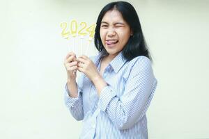 excited asian young business woman smiling with one eye blink holding golden candles numbers 2024 to celebrating new years eve wear blue stripes shirt looking at camera isolated on whtie background photo