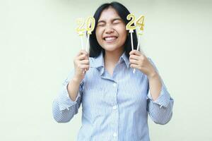 young business woman asian excited to celebrate new years eve by holding golden candles numbers 2024 with closed eye wear blu stripes shirt isolated on white background photo