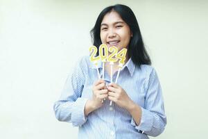 happy asian young woman office worker excited to celebrate new years eve by holding golden candles numbers 2024 wearing blue stripes shirt looking to camera isolated on white background photo