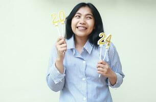 excited asian young woman laughing happy celebrating new years eve by holding golden candles numbers 2024 thinking resolution idea wearing blue stripes shirt looking away isolated on white background photo