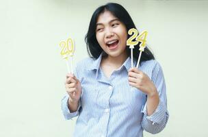 portrait of happy asian young woman coworker laughing excited to celebrate 2024 new year eve holding golden candles numbers wearing blue stripes shirt casual looking away isolated by white background photo