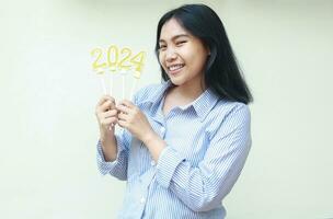 portrait of excited asian young business woman holding golden candles numbers 2024 for new years eve celebration wearing strip blue shirt looking to camera isolated on white background photo