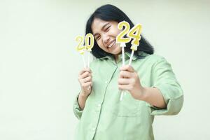beautiful and excited young woman asian smiling at camera showing 2024 number gold candles to celebrating new years eve wear green over size shirt isolated by white background photo