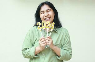 cheerful asian young asian female office worker excited to celebrate 2024 new years eve by holding gold number candles smiling to camera wear green over size shirt isolated on white background photo