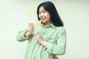 excited asian young business woman smiling at camera by holding gold glittering 2024 candles for new years eve celebration wearing green over size shirt isolated on white background photo