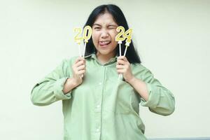 excited beautiful asian young woman blink eyes flirting at camera enjoy 2024 new years celebration by holding gold glittering number candles wearing green over size shirt isolated on white background photo