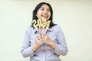 portrait of smiling asian young business woman excited for 2024 new years celebration ny holding gold figure candles on hand wearing grey formal suit looking away isolated on white background photo
