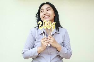 happy asian young woman smiling and wishing new resolution in 2024 new years eve celebration with holding number gold candles on hand wearing formal suit looking side isolated on white photo