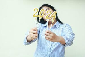 happy asian young business woman smiling and holding golden candles number 2024 covering face wear blue stripes shirt isolated on white background. beautiful female coworker celebrate new years eve photo