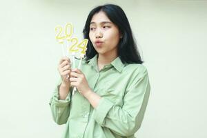 happy asian young business woman looking aside to golden candles number 2024 hold on her hand with duck face wear green over size shirt isolated white background. celebrating new years eve concept photo
