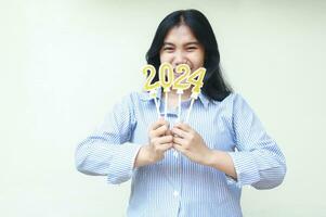 happy asian beautiful young woman holding golden candles number 2024 covering mouth to announce new years eve wearing blue stripes casual shirt looking to camera isolated over white background. photo