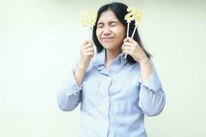 happy and excited asian young woman holding golden candles number 2024 stick on her head with eyes closed to celebrate new years eve wear blu stripes shirt isolated on white photo