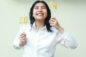 excited asian young woman coworker smiling and looking aside to number 24 of 2024 figure candles hold on lifted hand wear white shirt, isolated, female wish new resolution in new years eve photo