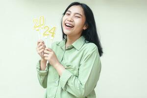 happy asian young woman wearing casual green shirt laughing at camera with lifting arm holding 2024 figure candles isolated on white, female celebrating new years eve photo