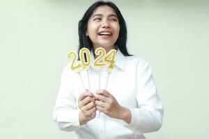 carefree asian young woman smiling and looking aside to copy space while showing 2024 figure candles on hand to announce new years eve wearing white formal shirt, isolated photo