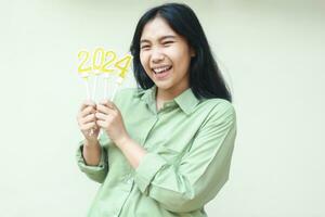 cheerful asian young woman smiling to camera wearing green casual clothes with lifitng arm holding 2024 figure candles to celebrate new years eve, isolated over white background photo