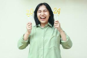 portrait of excited asian young woman hipster laughing with closed eye and rasing arm with 2024 figure candles hold on hand wearing green over size shirt, isolated on white. new years eve concept photo