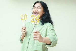 portrait of carefree asian young woman laughing at camera with closed eye and show 2024 figure candles lifting on hand wearing green over size shir isolated on white background. looking to camera photo