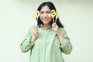 happy asian young woman holding golden candles 2024 number and looking at camera with smiling face wear grean over size shirt celebrating new years eve isolated on white background photo