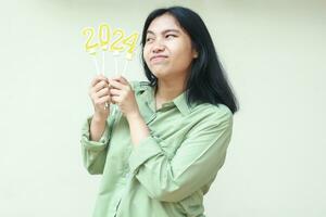 happy asian young woman looking aside to glittering 2024 number candles think resolution in new years celebration wear over size green shirt, isolated on white background photo