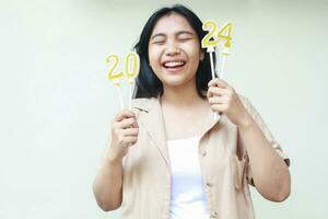 attractive asian young woman laughing with eye closed and raising arm holding 2024 golden candles wearing casual brown outer celebrating new years eve isolated on white background photo