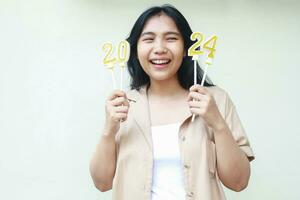 carefree asian young woman laughing and looking aside with holding 2024 candles figure wearing casual brown outer to celebrate new years eve isolated over white background photo