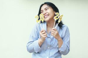 ecstatic asian young business woman wear blue stripes casual shirt holding 2024 figure candles looking aside to copy space with smiling face excited to celebrate new years eve, isolated on white photo