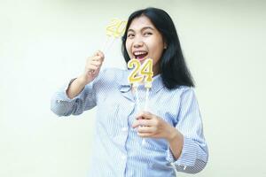 excited asian young business woman wear blue stripes stylish clothes with arm lifted holding 2024 figure candles for new years eve celebration, female smiling and looking away photo