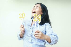 alegre asiático joven mujer celebrando nuevo años víspera con risa y contento sonrisa, mirando arriba con cerrado ojo, hembra levantamiento brazo sostener 2024 figura velas vestir azul rayas de moda ropa aislado foto