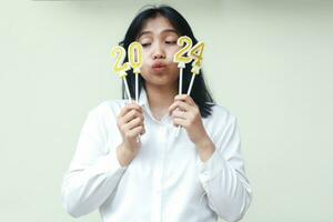 thoughtful asian young woman holding 2024 figure candle with curiosity and looking aside to number 20 wearing white formal shirt, isolated photo