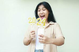 excited asian young woman hipster wear fashionable brown casual outer screaming and looking to 2024 number candle hold on hand, isolated on white, beautiful female enjoy new years eve celebration photo