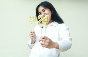 happy asian young business woman wearing white formal shirt excited to announce new years eve by showing number 24 of 2024 number candle hold on hand, looking at camera with eye closed, isolated photo
