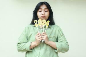 thoughtful asian young woman thinking and looking down to 2024 number candles holding by hand wearing green over size shirt, new years eve concept, isolated on white background photo