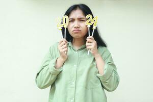 confused asian young woman raising 2024 number candle to her face by hand, beautiful female wearing green over size shirt, looking to camera, new years eve concept, isolated over white background photo
