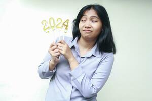 happy young asian woman thinking about 2024 figure candle hold on her hand while looking aside wearing grey formal shirt, isolated on white background photo