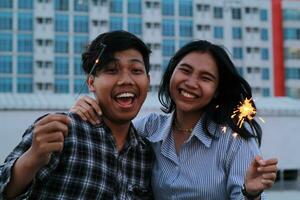 carefree asian teen couple holding sparkling firework and dancing celebrating new year with laugh wear flannel and stripes shirt over urban building background photo