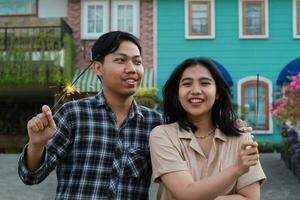 candid asian couple enjoying new year celebration by holding sparkler firework in the house yard green garden wearing casual outer and flannel photo