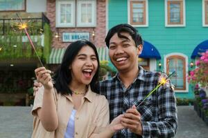 exited asian young couple holding sparkler in the colorful house yard green garden wear stylish clothes look to camera, friend screaming celebrate new year photo