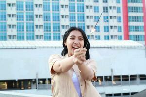 beautiful asian young woman holding sparklers firework and laughing in new year eve celebration standing in roof top outdoor with urban building background photo