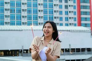 excited asian young woman holding sparkler to celebrating new year eve in rooftop apartment with city building background photo