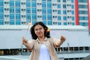 contento asiático joven mujer hipster participación bengalas Fuegos artificiales celebrar nuevo año víspera con bailando en techo parte superior Departamento con urbano edificio antecedentes foto